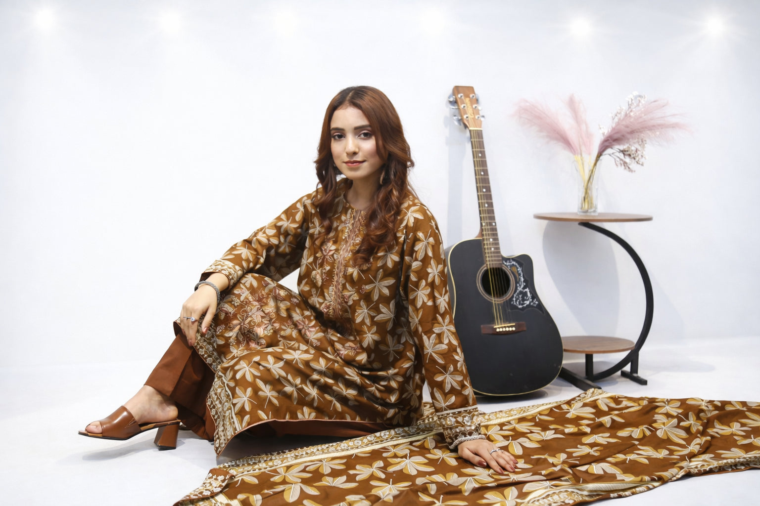 Woman in a patterned outfit sitting on the floor with a guitar and decorative items in the background.