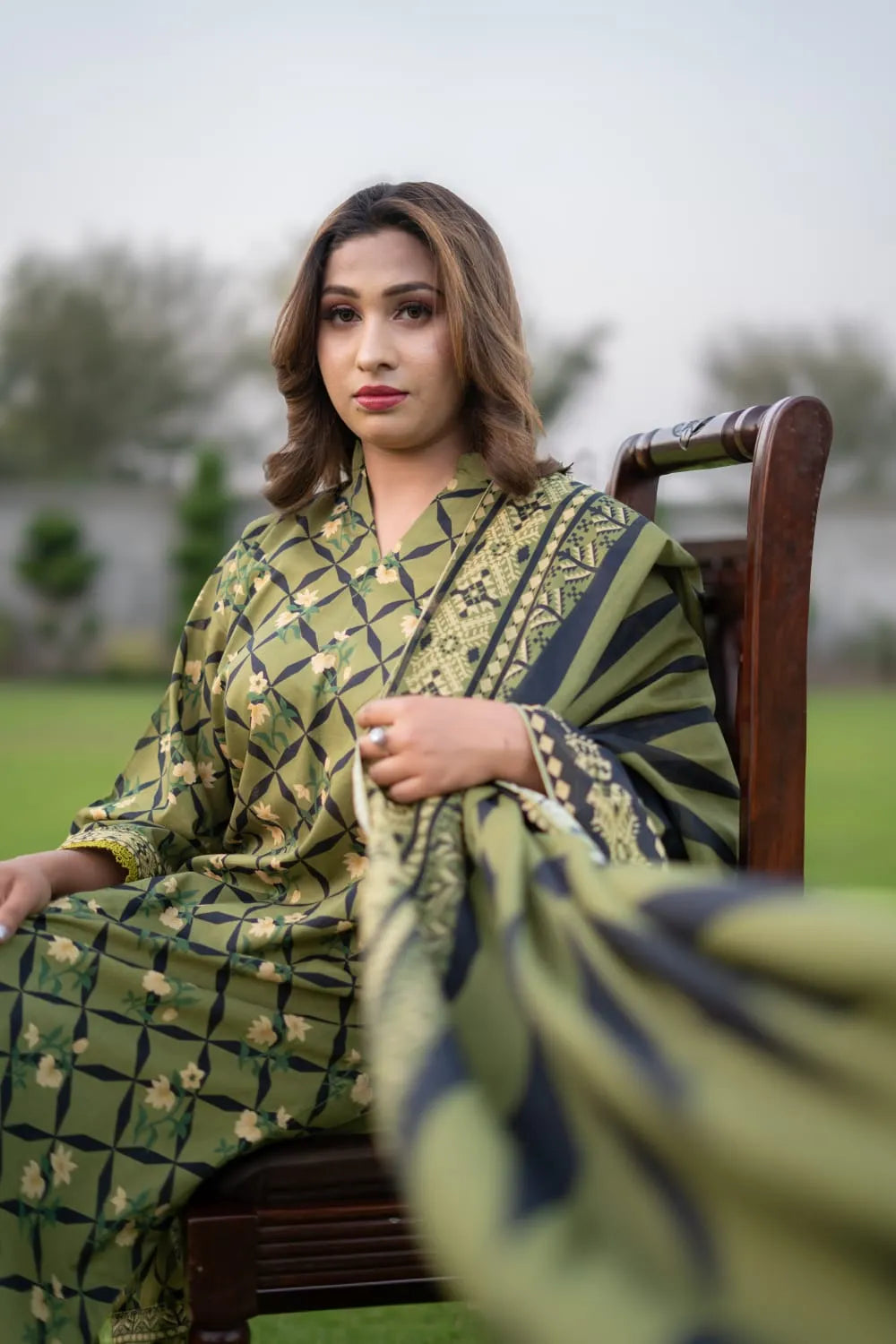 Woman in a green patterned outfit sitting on a wooden chair outdoors.