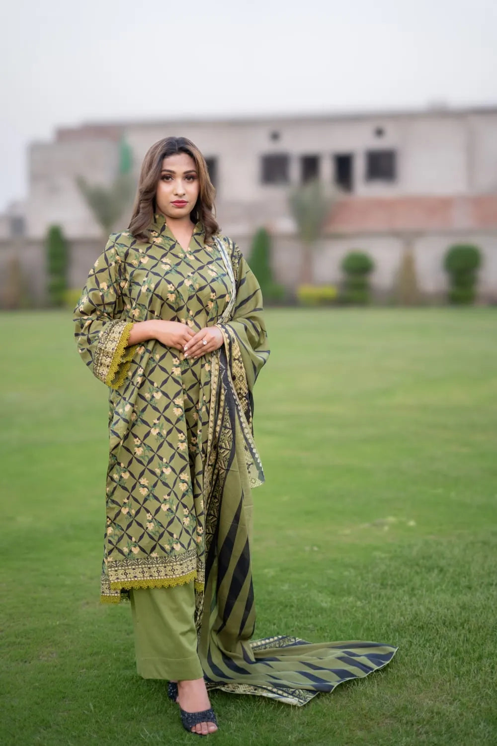 Woman in a green and black traditional outfit standing on grass with a building in the background
