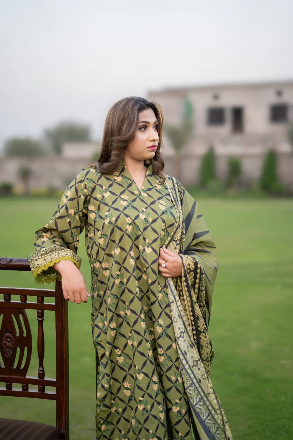 Woman in a green and gold traditional outfit standing outdoors on a grassy area.