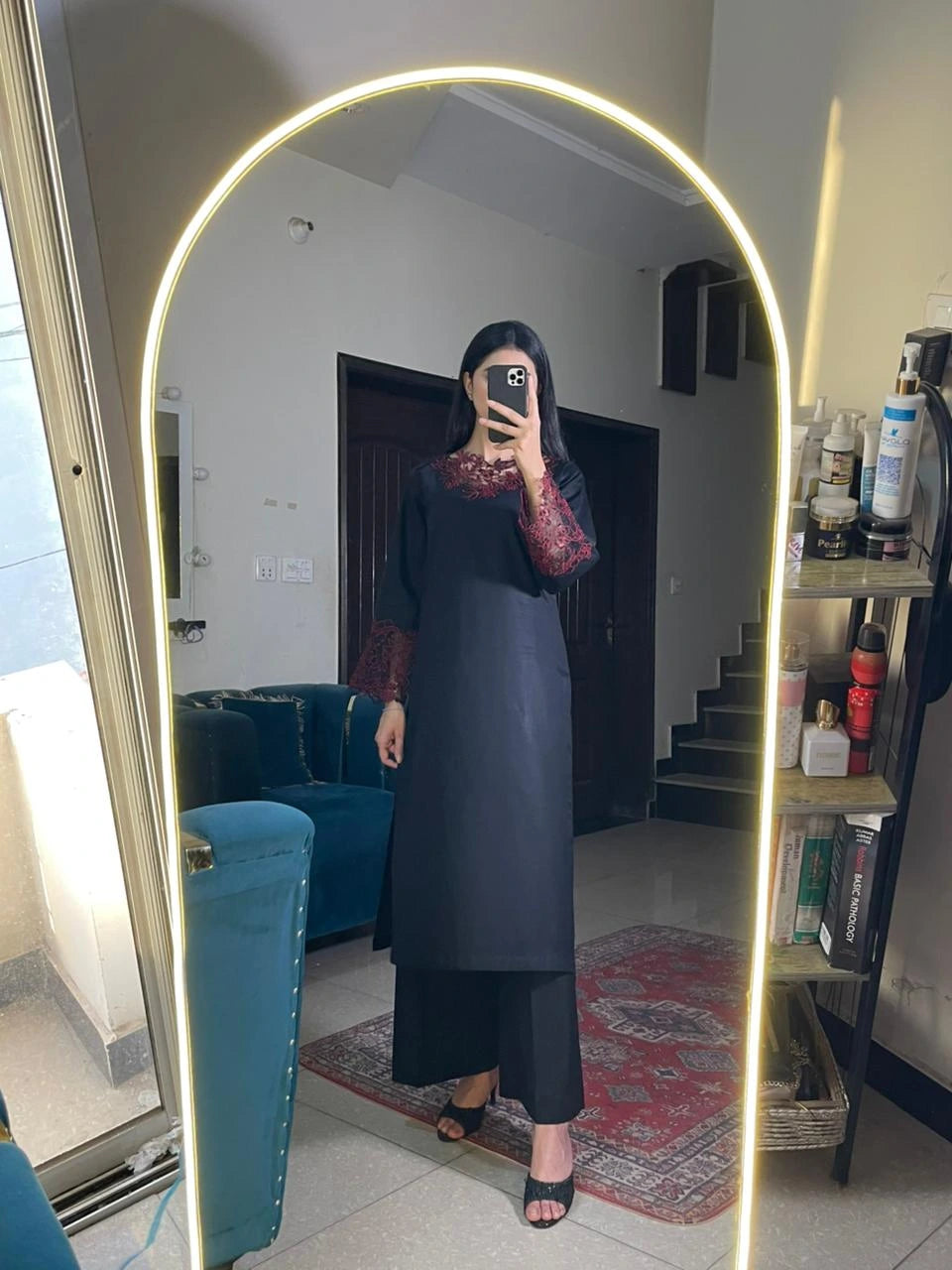Person taking a mirror selfie wearing a dark dress in a room with a blue couch and bookshelf.