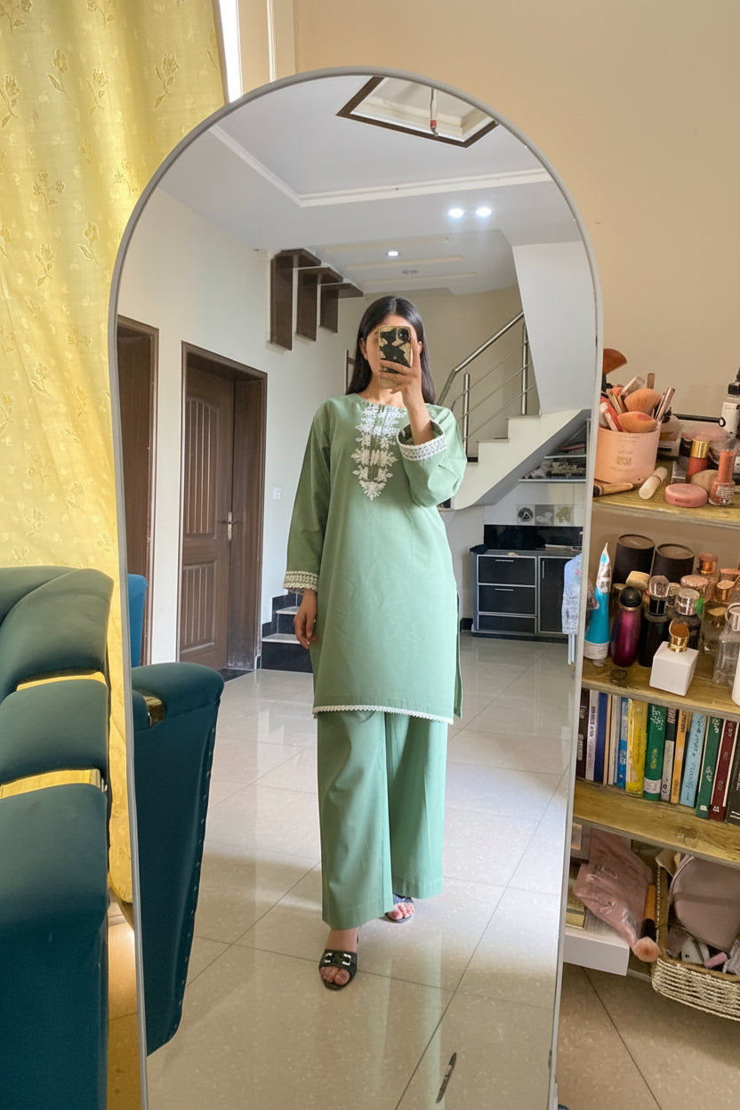 Person wearing a green outfit taking a mirror selfie in a room with a bookshelf and furniture.