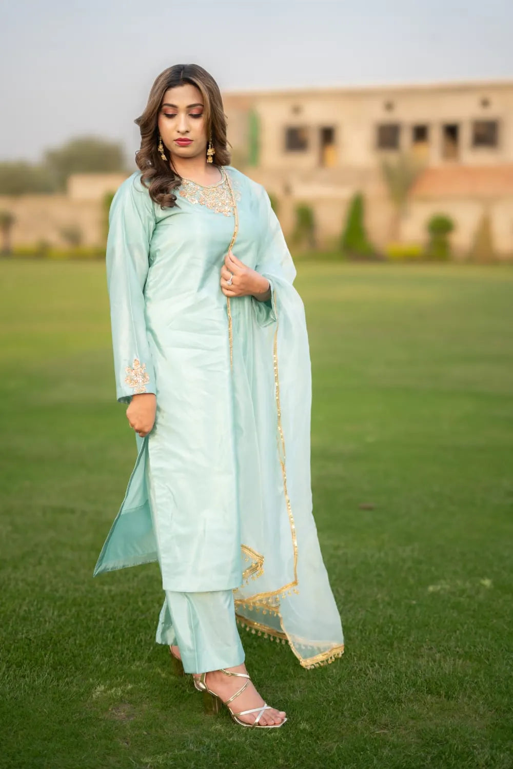 Woman in a light blue traditional outfit standing on grass with a blurred building in the background