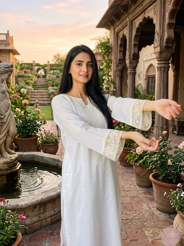 Woman in a white traditional outfit standing in an outdoor setting with architectural elements and plants.
