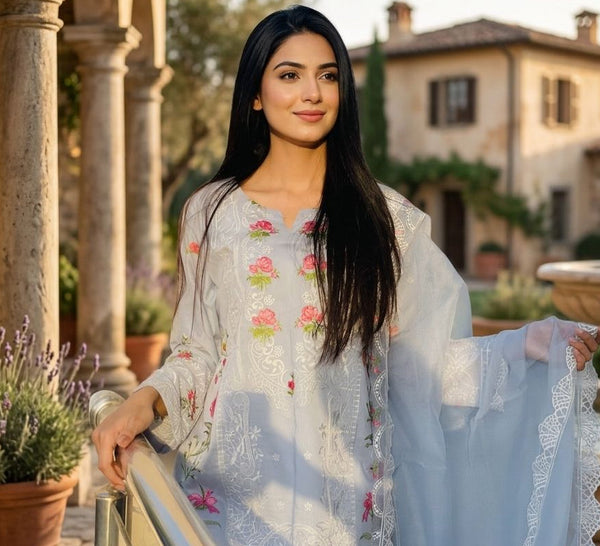 Woman in a light blue embroidered outfit standing in an outdoor setting with columns and a fountain.